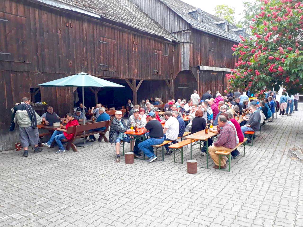 Eine Menschgruppe sitz nach dem Himmelfahrtsmarsch an Biertischen in einem Hof eines Bauernhofs unter blühenden Bäumen bei Getränken und Grillwürsten