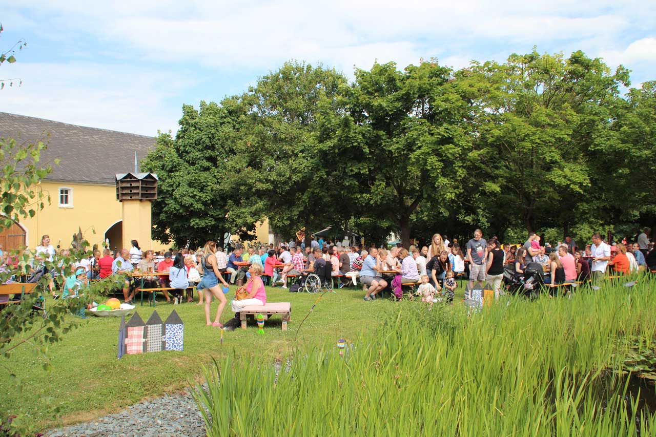 Sommerfest Gut Münchenreuth: eine fröhlich im Sonnenschein an Biertischen feiernde Menschenmenge auf einer Wiese vor dem Gut Münchenreuth